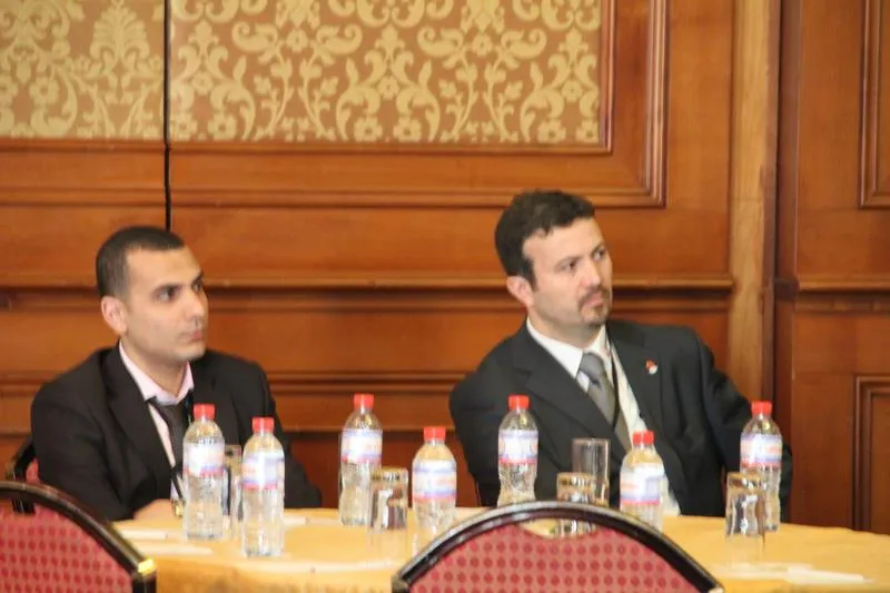Walid and Malik Saheb, both in suits, seated side by side at a table with water bottles, listening to a talk
