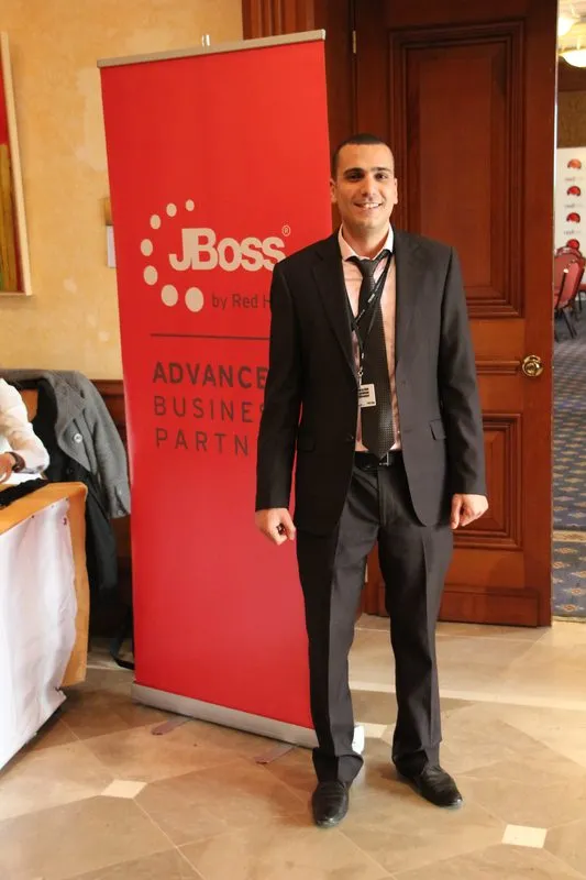 A man in a dark suit and lanyard standing beside a red "JBoss by Red Hat Advanced Business Partner" roll-up banner