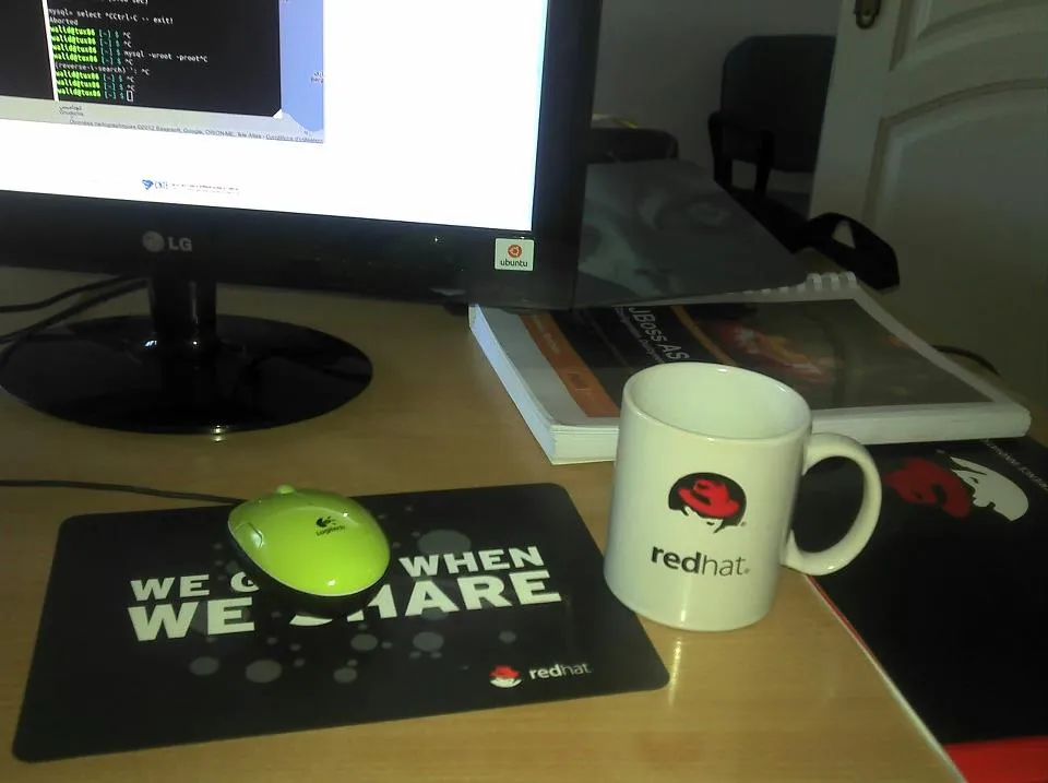 A white Red Hat mug and a black Red Hat mouse pad reading "We grow when we share" on a desk, next to a JBoss AS book and a monitor running Ubuntu